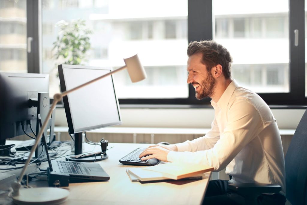 man-in-white-dress-shirt-sitting-on-black-rolling-chair-while-facing-black-computer-set-and-smiling-840996 A man smiling while working at an office desk with a computer and natural daylight streaming in through large windows.