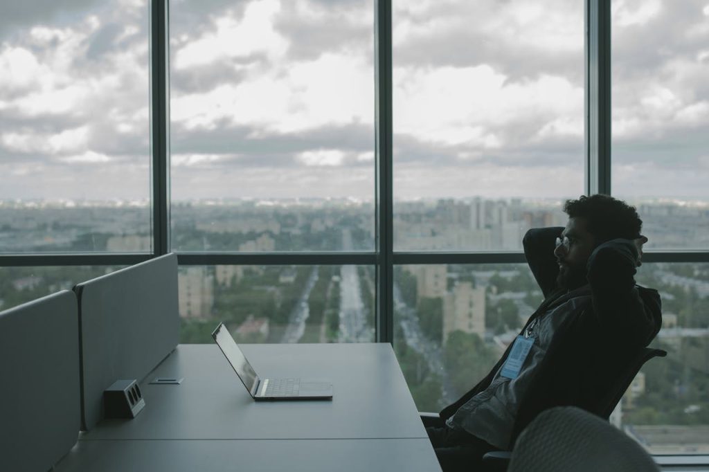 man-reclining-and-looking-at-his-laptop-5483064 A man sits in an office with hands on head in front of a laptop, overlooking a cityscape.